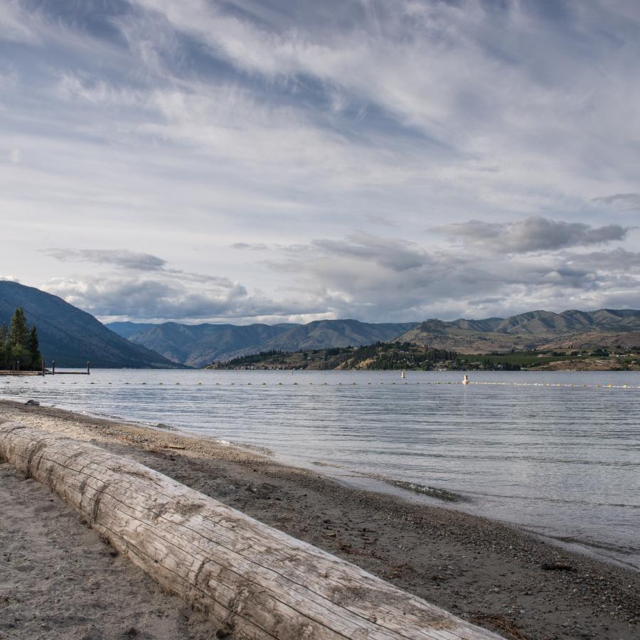 A gravel beach with a log berm in front of the calm waters of Lake Chelan which are reflecting a cloudy sky and the distant mountains.