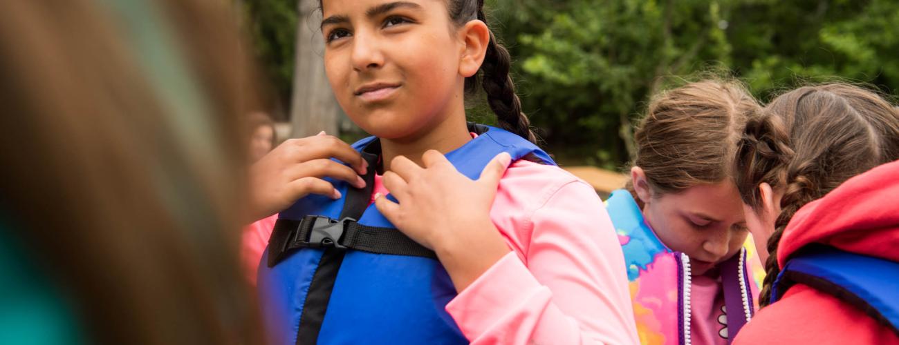 A young girl wearing a life jacket. Others in the background are putting life jackets on.