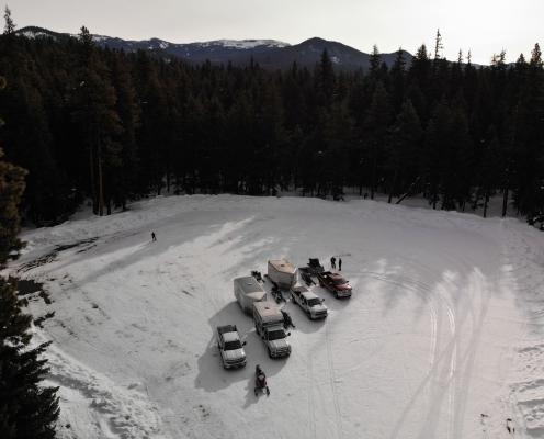Four trucks, two with trailers, and five snowmobiles stand in the middle of a snow-covered parking area surrounded by a thick pine forest.