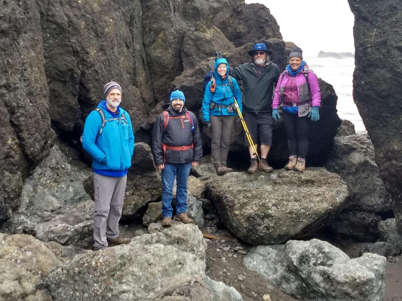 Five people wearing rain jackets stand on rocks with the ocean in the background
