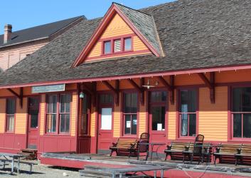 Old western style building painted orange with red trim with a sign on the left front reading, "Cle Elum". Wooden park benches sit on the front, open porch of the building. Picnic tables sit on the dirt in front of the building.