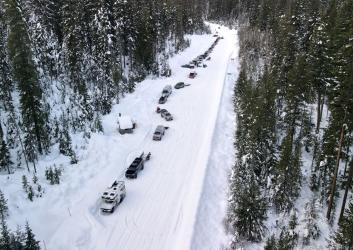 Trucks prarallel-parked along the side of a forest road covered by thick snow and flanked by snow-dusted pine tree forest. A toilet pit hut lies on the edge of the tree line.