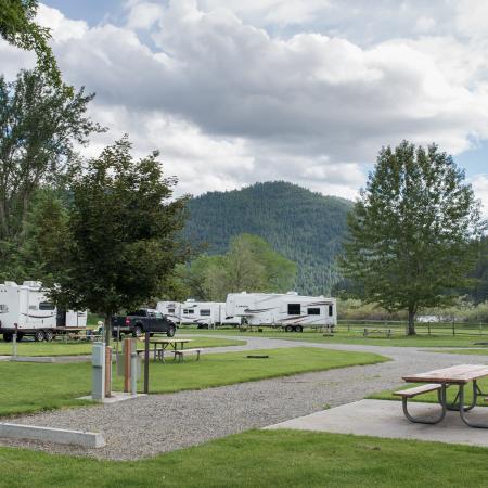 Gravel RV and trailer campsites with hook-ups and picnic tables are shown with a few large camping trailers, tall trees and mountains in the background.
