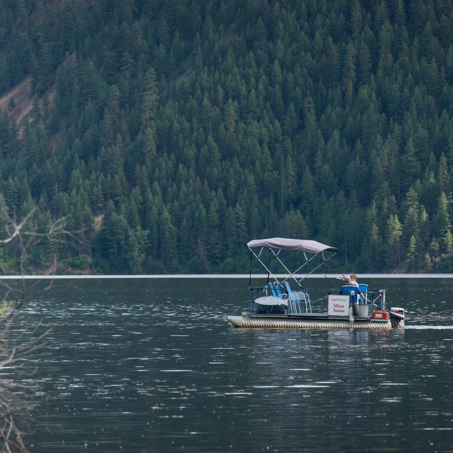 Two people on a motorized boat on a reservoir with a dam in the background and a hillside covered in trees on the far side of the water.