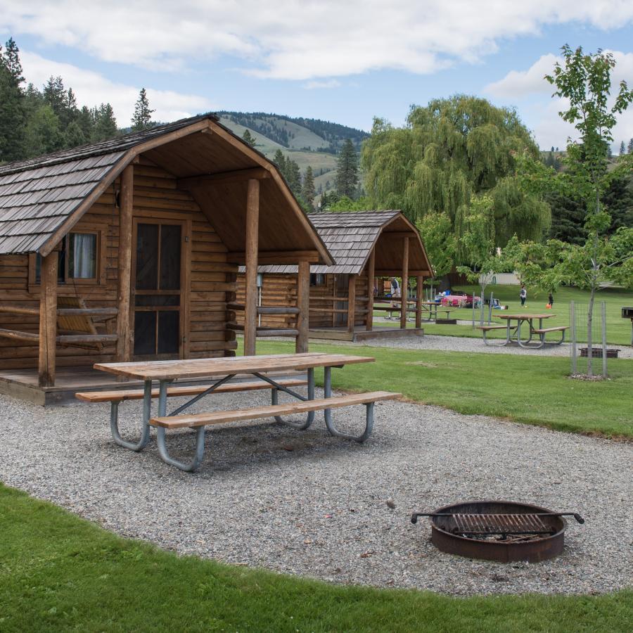 Two log cabins with picnic tables, fire rings, and barbeque pits with trees and hills in the background.