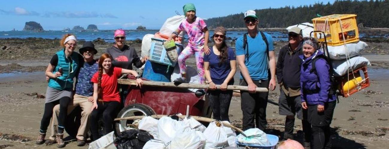 Nine volunteers standing on the beach around garbage in bags, broken buoys, a tire and more that was collected. A blue sky, ocean and small islands are seen in the background.
