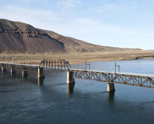 A n aerial shot of the Beverly Bridge, which spans the gray-blue waters of the Columbia River. A dark-tan, high-desert hilllside rises toward a pale blue sky in the background.