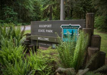 Rockport State Park wooden entrance sign with the state parks shield and the word "welcome." In front of the sign is ferns and in the background lush green trees and undergrowth. 