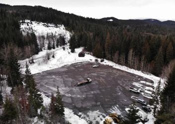 Large square paved parking lot plowed clean surrouned by pine trees next to a forest road with light snow. A warming hut and pit toilet hut stand in the upper right corner and several trucks are parked along the right edge of the lot.