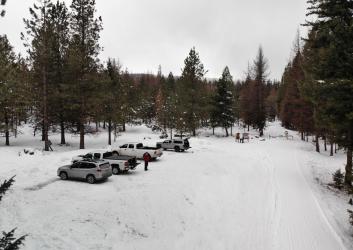An unplowed parking area with compacted thin snow at a trailhead, next to a groomed road, surrounded by snow-dusted trees.