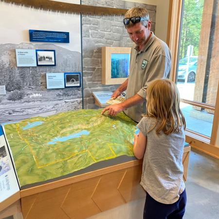 A park staff employee wearing a grey polo shirt with the State Park logo talking about a topographic map of the area with a girl with blonde hair and a grey shirt.