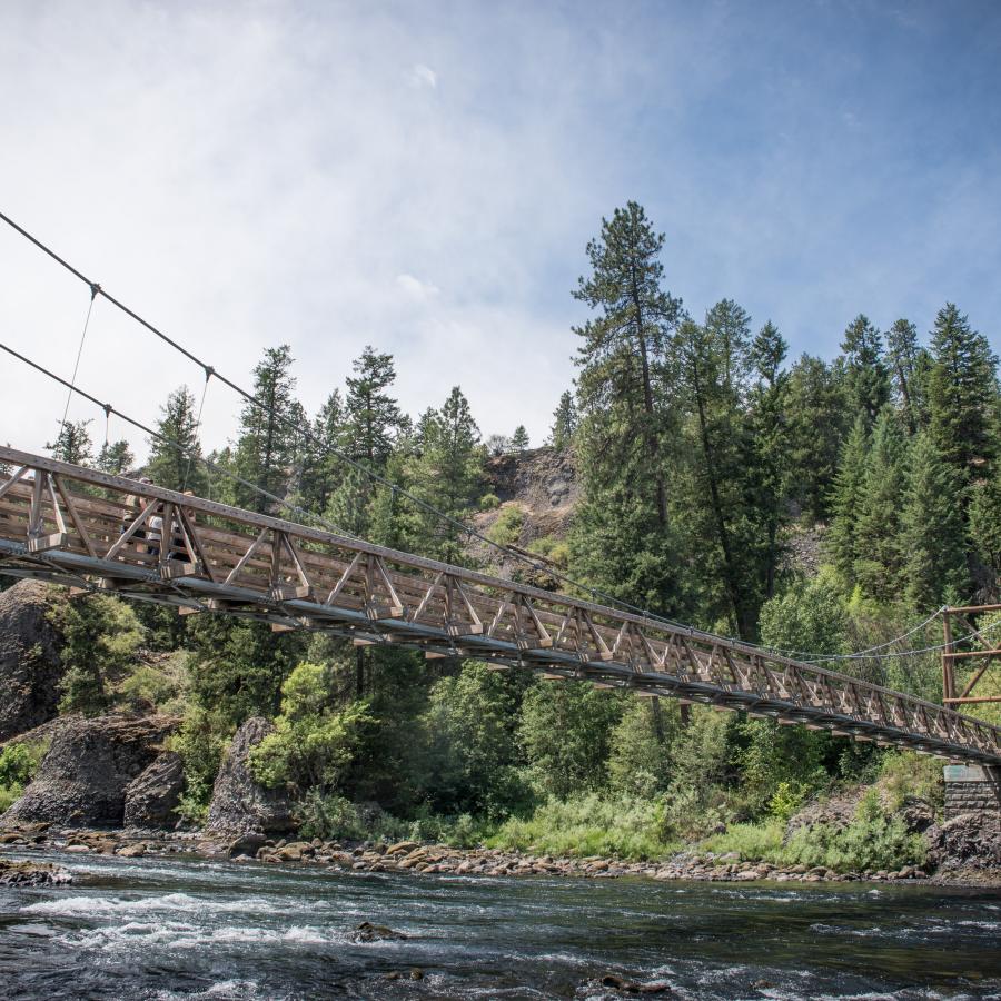 A suspension bridge spans the Spokane River with trees covering the far bank and partly cloudy skies.