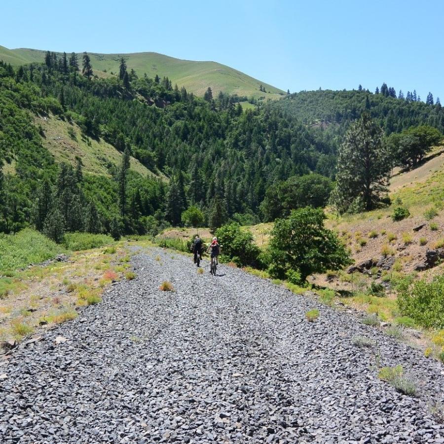 Two cyclists ride on a wide gravel trail with tree covered hillsides on either side.