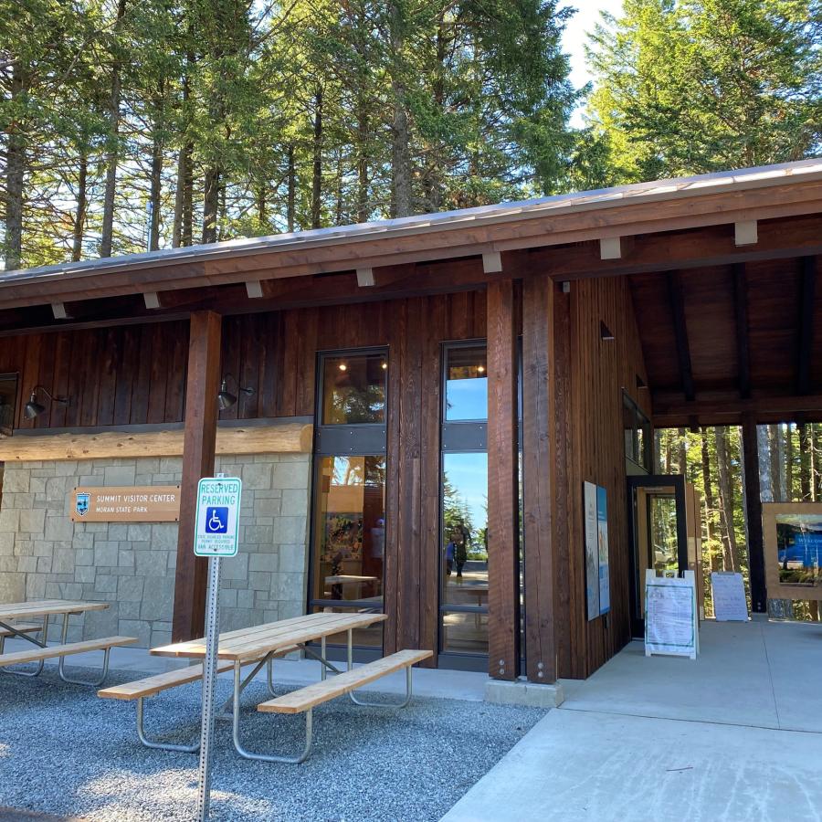 An exterior view of the visitor center with dark brown wood supports and siding, tall floor to ceiling windows and wall of rock. Picnic tables sit next to the building on pea gravel. 