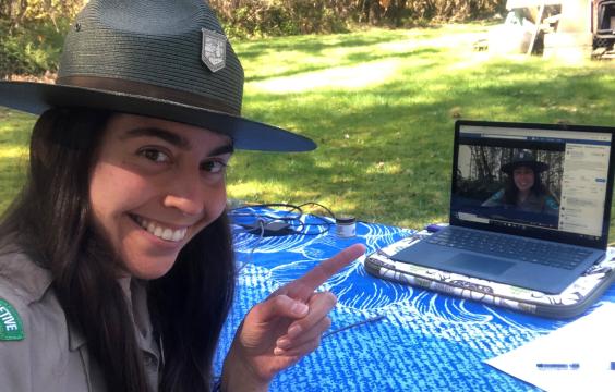 A person in a Washington State Parks ranger uniform points to a laptop screen on a table. On the laptop is a video of the same ranger conducting a virtual interpretive program.